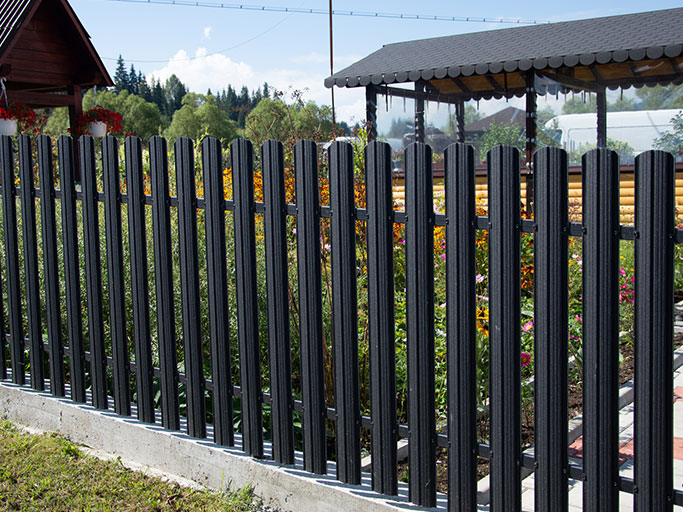 Black vertical metal fence bordering a colorful flower garden with a wooden gazebo and trees in the background