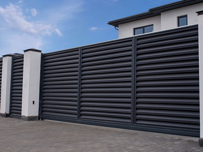 Modern dark horizontal slat metal driveway gate and white stucco pillars in front of contemporary house under blue sky