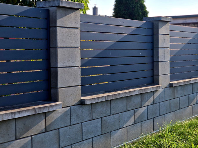Modern gray concrete block fence with horizontal slats and pillars casting diagonal shadows over lawn