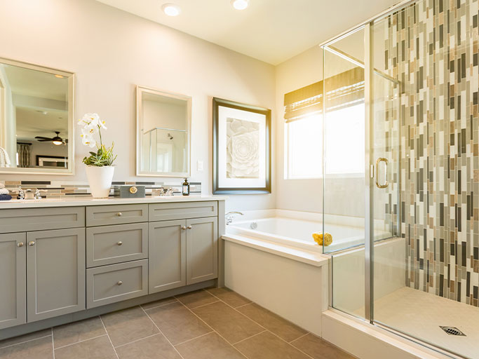 Bright modern bathroom with gray double-sink vanity, soaking tub, glass shower with vertical mosaic tile and window light