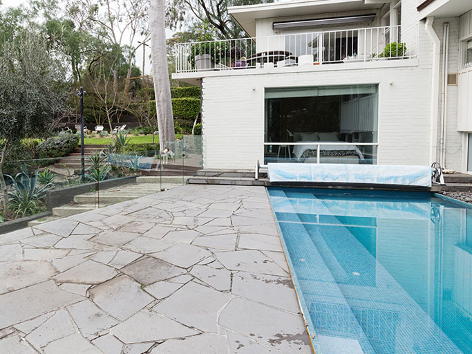 Modern backyard with rectangular swimming pool, stone patio, glass pool fence and white two-story house with balcony.