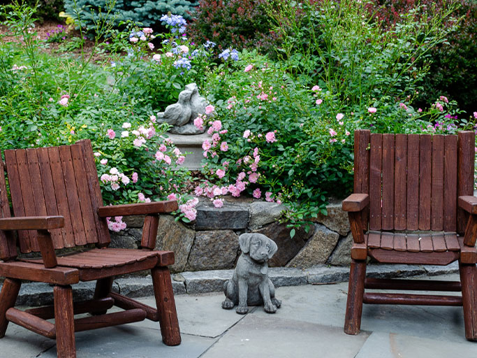 Two wooden Adirondack chairs flanking a small stone dog statue in a rose-filled garden with a stone retaining wall