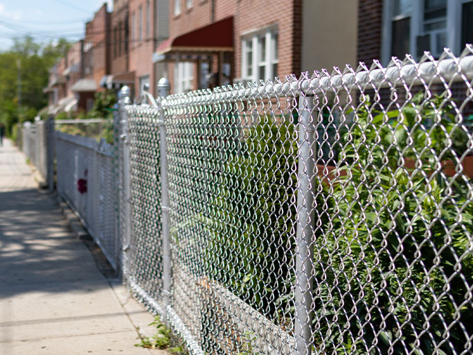 Chain-link fence along a residential sidewalk with brick row houses and green shrubs