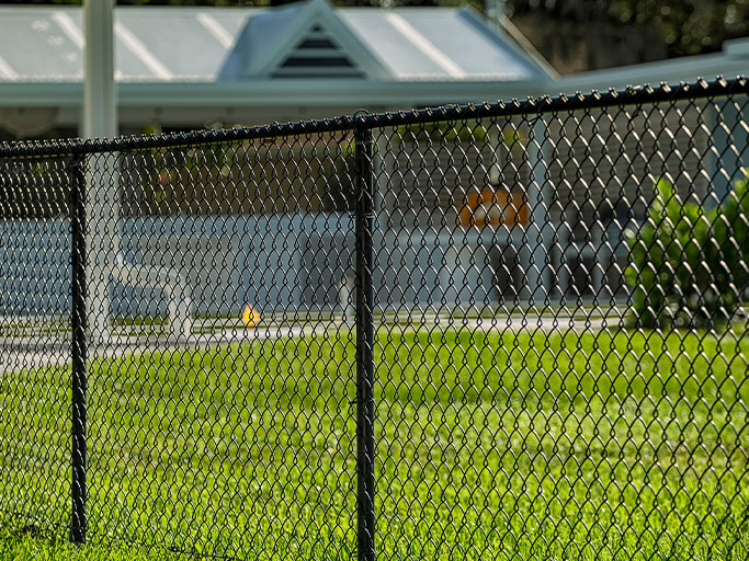 Black chain-link fence in foreground with sunlit green lawn and pool area under a covered pavilion