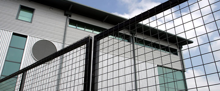 Modern industrial building with corrugated metal siding, teal windows and round vent behind a black wire fence, angled view