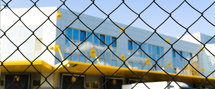 Chain-link fence foreground, modern industrial building with blue windows and yellow accents in background