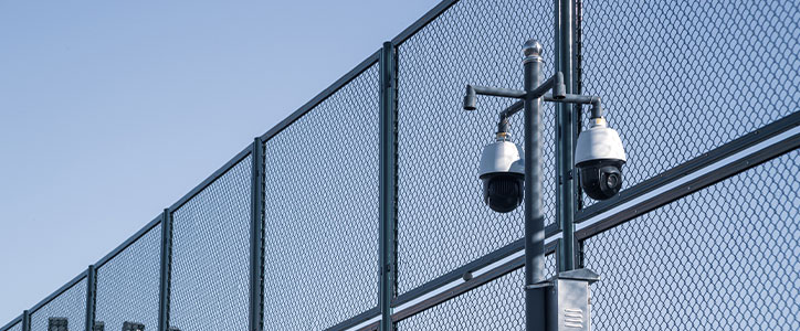 Two dome security cameras on a pole beside a tall chain-link fence against a clear blue sky