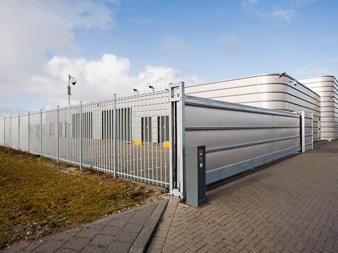 Modern industrial facility with metal security fence and sliding gate, surveillance camera and paved driveway.