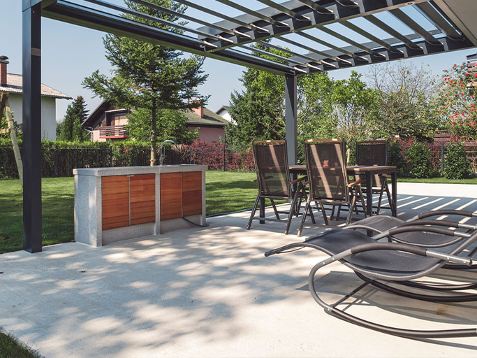 Modern backyard patio with pergola, outdoor dining set, built-in counter and lounge chairs overlooking landscaped lawn.