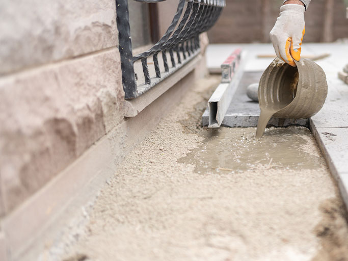 Worker pours wet concrete into joint between patio pavers along house foundation.