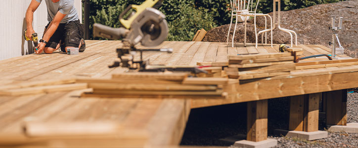 A man installing wooden deck boards using a drill and circular saw on a backyard deck construction site.