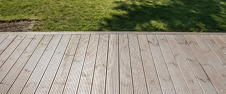 Sunlit wooden deck boards in foreground with green lawn and tree shadows in background, outdoor patio yard.