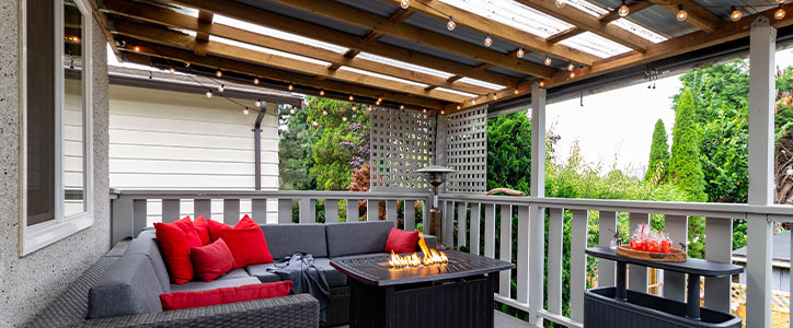 Cozy covered patio with gray wicker sectional, red pillows, fire-pit coffee table, string lights and garden view