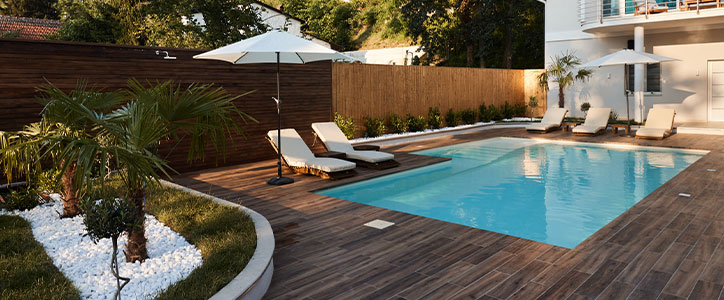 Modern backyard swimming pool with wood deck, sun loungers and umbrellas, palm plants and white stone landscaping.