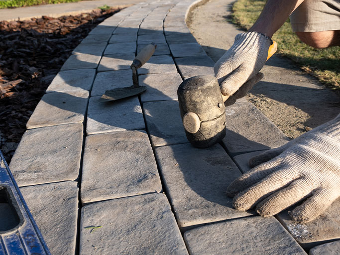A person in gloves using a rubber mallet and trowel to install gray stone pavers on a curved walkway.
