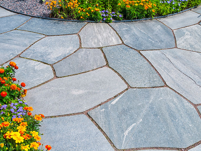 Curved flagstone patio with irregular gray pavers and colorful marigolds and purple flowers along edge.