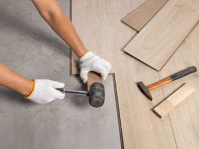 A person wearing gloves taps a laminate plank into place with a rubber mallet and tapping block.