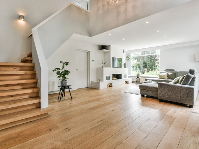 Open-plan modern living room with wide oak floors, wooden staircase, gray sectional sofa, fireplace and large window.