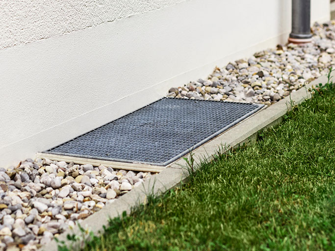 Metal basement window well cover set in gravel beside house wall with adjacent lawn and downspout