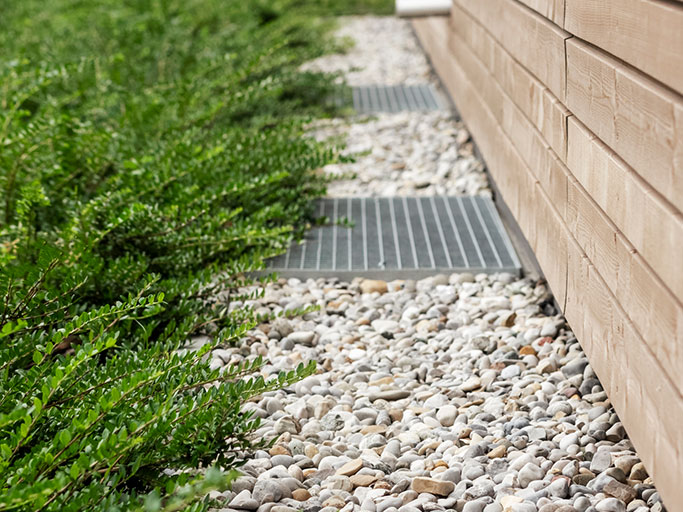 Gravel drainage bed with white pebbles, metal grates, green groundcover and wood siding along a house foundation