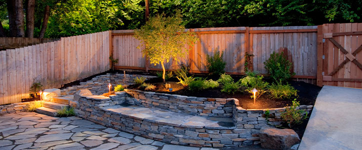 Evening backyard with stone patio, terraced retaining walls, planted garden beds and warm landscape lighting by wooden fence