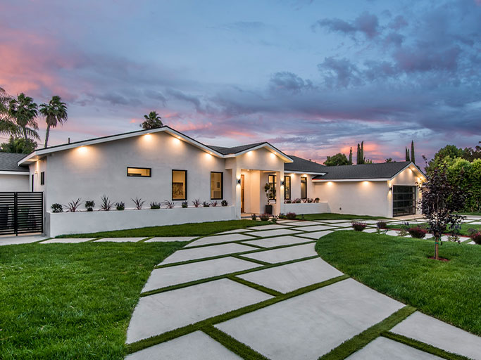 Modern single-story home at dusk with illuminated exterior, landscaped lawn and geometric concrete driveway