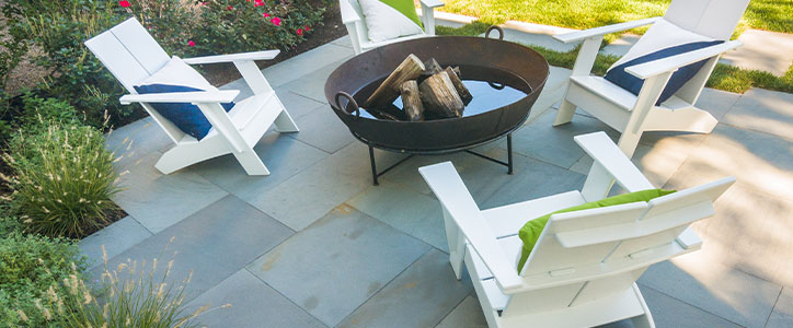 Four white Adirondack chairs with blue and green cushions arranged around a round fire pit on a stone patio