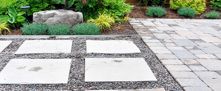 Modern backyard patio with large square concrete stepping stones set in gravel beside stone pavers and landscaped plants.