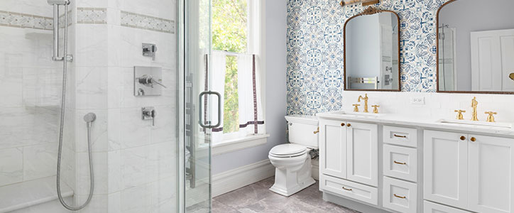 Bright modern bathroom with white double vanity, gold fixtures, walk-in glass shower, patterned blue accent wall and window.