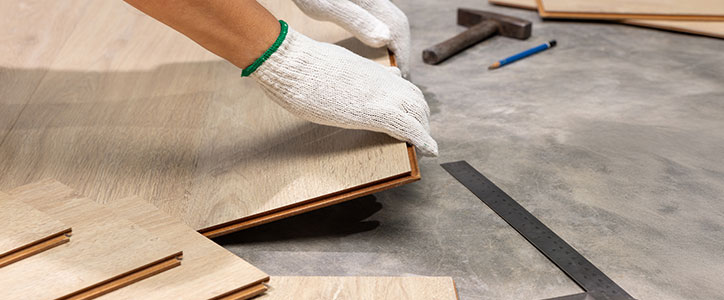 A person wearing gloves installing interlocking laminate flooring planks on a subfloor with measuring tools nearby