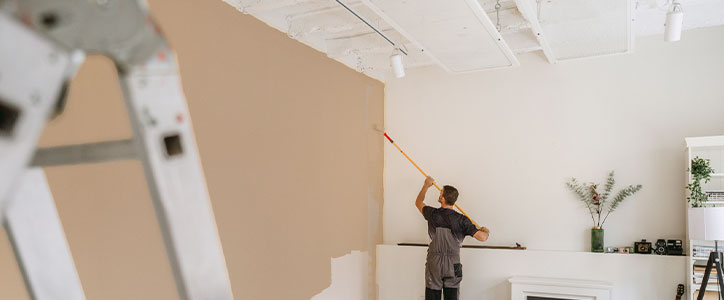 A man painting a living room wall beige with an extended roller, ladder in foreground and decor on mantel.