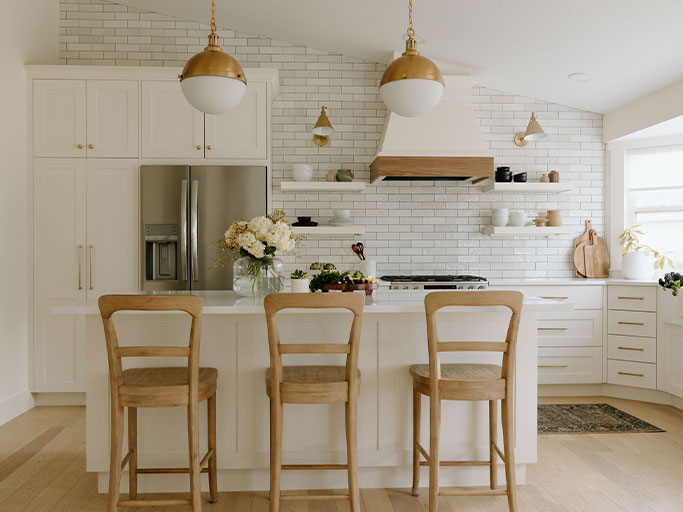 Bright modern white kitchen with island, three wooden stools, brass pendants, subway tile backsplash, stainless fridge