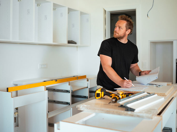 A man reviewing kitchen renovation plans and taking measurements among cabinets, level and power tools on the countertop.