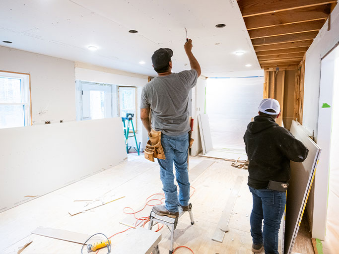 Two workers installing drywall during home renovation; one on step stool fastening ceiling panel, another holding drywall.