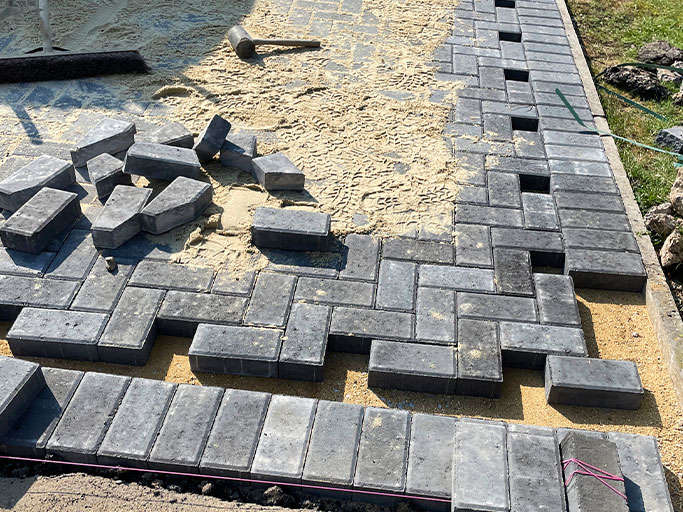 Gray interlocking concrete pavers being installed on sand base with scattered blocks and mallet