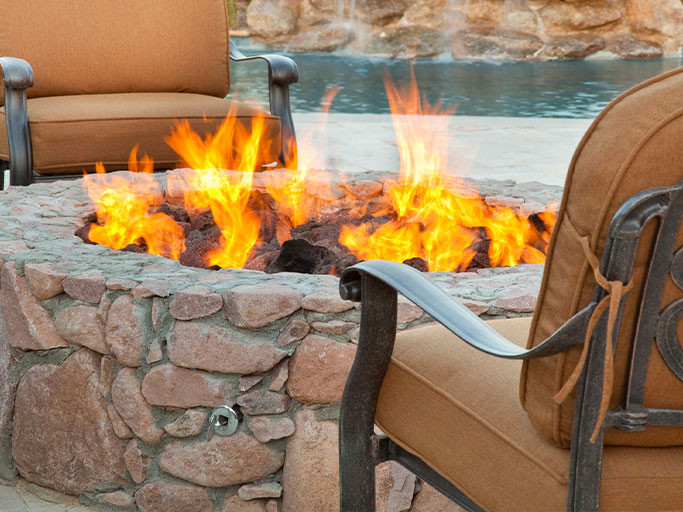 Stone patio fire pit with bright orange flames, surrounded by cushioned outdoor chairs beside a pool.