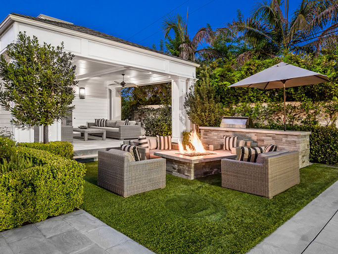 Outdoor patio with wicker seating around a lit fire pit, built-in grill, covered lounge and umbrella on manicured lawn