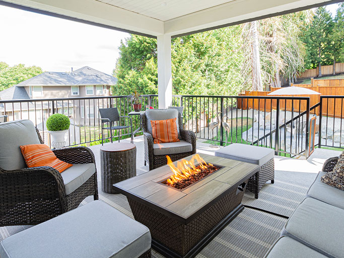 Covered patio with wicker seating and orange pillows around a rectangular fire pit table, overlooking a fenced backyard