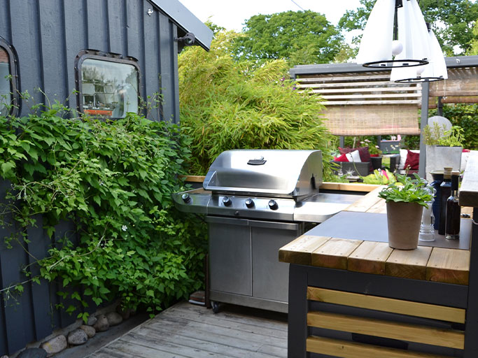 Outdoor patio kitchen with stainless steel gas grill, wooden counter, potted plant and lush privacy greenery.