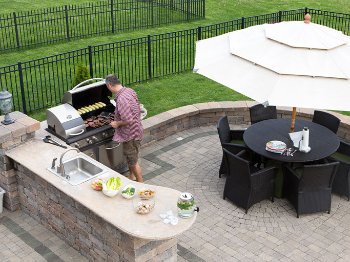 A man grilling at a backyard outdoor kitchen with stone counter, sink, bowls of salad, and umbrella-shaded dining table