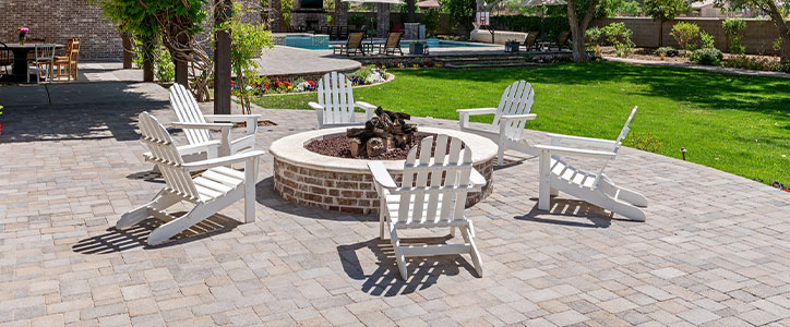 Backyard paver patio with white Adirondack chairs arranged around a circular brick fire pit, lawn and pool in background