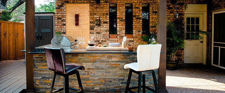 Outdoor patio kitchen with stone-front bar, built-in grill and two bar stools by brick wall.