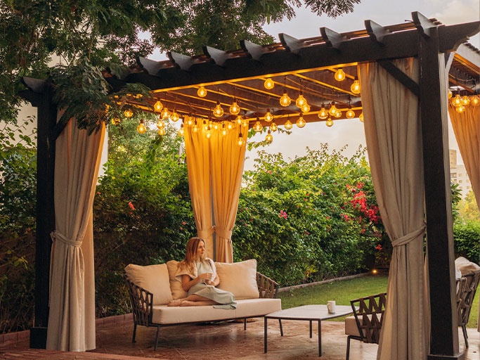 Woman relaxing on patio sofa under pergola with string lights, cozy evening garden seating area.