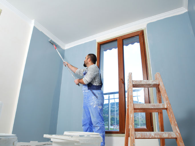 A man painting interior walls blue with a roller near the ceiling, ladder and window nearby.