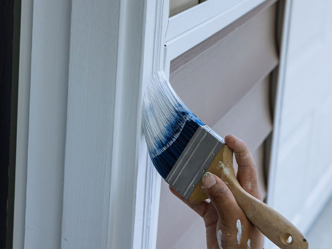 A person painting exterior house trim with a brush, applying white paint to window casing and siding.