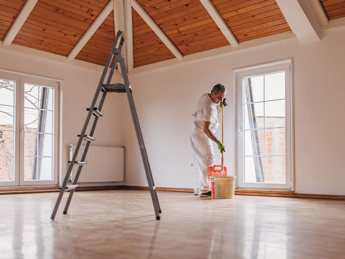 A man mopping a hardwood floor in an empty sunlit room with ladder, large windows and vaulted wooden ceiling.