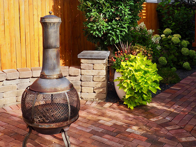Rustic metal chiminea on a brick paver patio with stone wall, wooden fence and lush potted plants in a backyard.