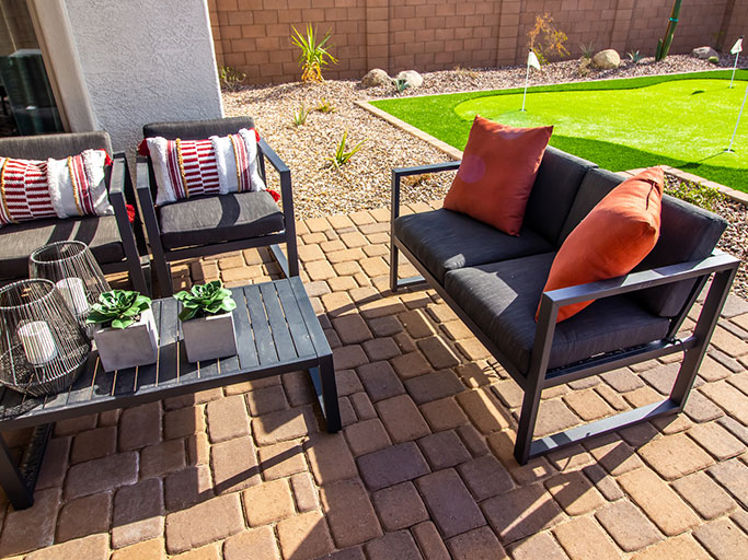 Patio seating on brick pavers: gray chairs and loveseat with orange pillows, coffee table with succulents, putting green