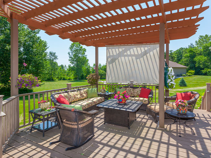 Sunlit backyard patio with wooden pergola, wicker seating, colorful cushions and potted flowers overlooking lawn.
