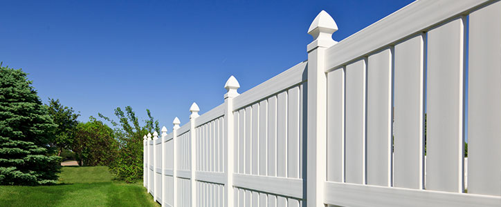 Long white vinyl privacy fence bordering a green lawn with trees under a clear blue sky